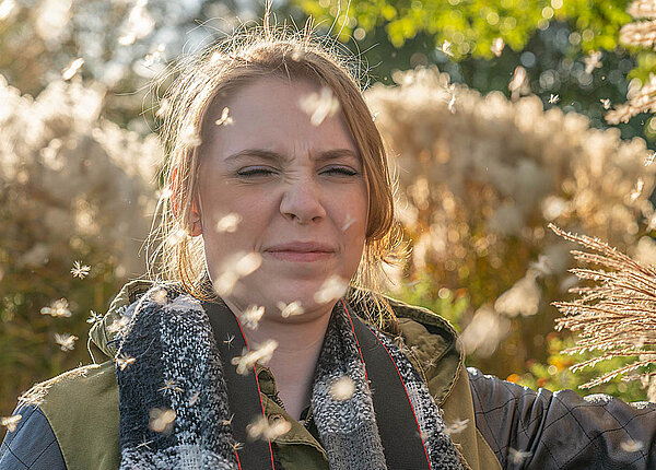 Allergie: Bei Heuschnupfen sind Pollen die Allergene. Eine Frau steht im Freien und Pappelpollen fliegen an ihrem Gesicht vorbei. Sie verkneift das Gesicht.