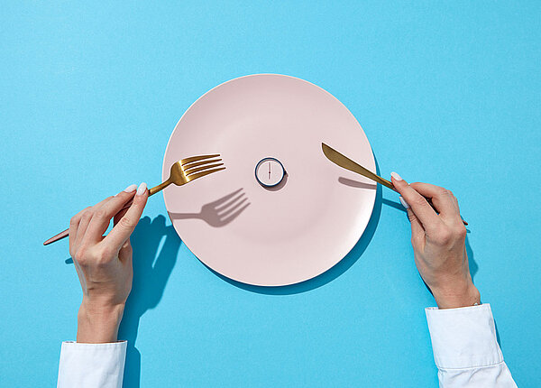 Fasten als Selbsttest Weißer Teller mit kleiner Uhr drauf, daneben hält Frau Messer und Gabel in den Händen
