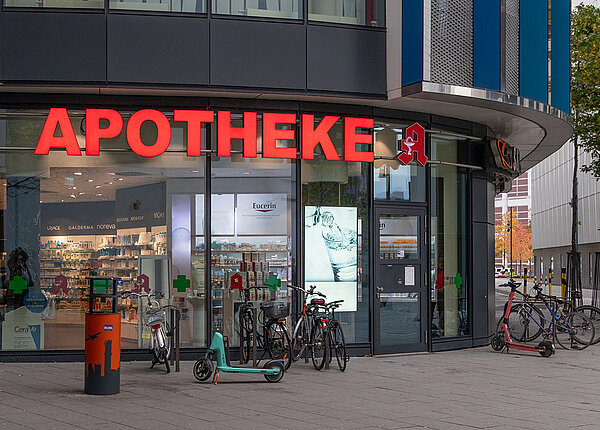 Apothekenvergütung und Apothekenreform Eingang einer Apotheke mit Glasfassade. Davor stehen Fahrräder und Roller.