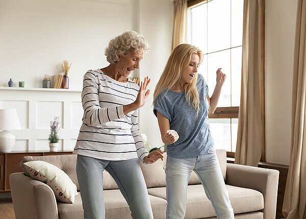 Inkontinenz Zwei Frauen, eine in den 30ern und eine in den 50ern, tanzen ausgelassen in einem Wohnzimmer vor dem Sofa