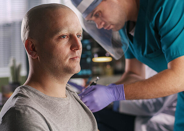 Verbessert die Corona-Impfung eine Immuntherapie gegen Krebs? Ein Krankenpfleger oder Arzt mit Gesichtsvisier impft einen Mann mit Glatze.