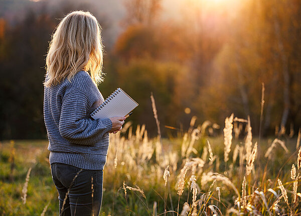 Motivation im Alltag steigern Rückansicht einer blondhaarigen Frau, die in der rechten Hand einen Block und Stift hält und sich den Sonnenuntergang anschaut.