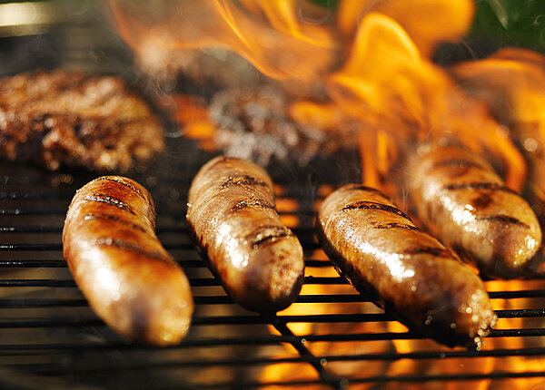Zu dunkle Würstchen Im Vordergrund vier Würstchen auf einem Grillrost, im Hintergrund ein paar Steaks mit Flammen