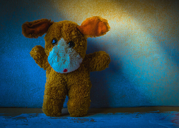 Schlafstörungen durch die Psyche: Ursachen und Therapie Ein Kuscheltier steht allein in einem dunklen Raum, nur erhellt von einem Lichtstrahl. Es hat große runde Augen und hebt die Arme nach oben.