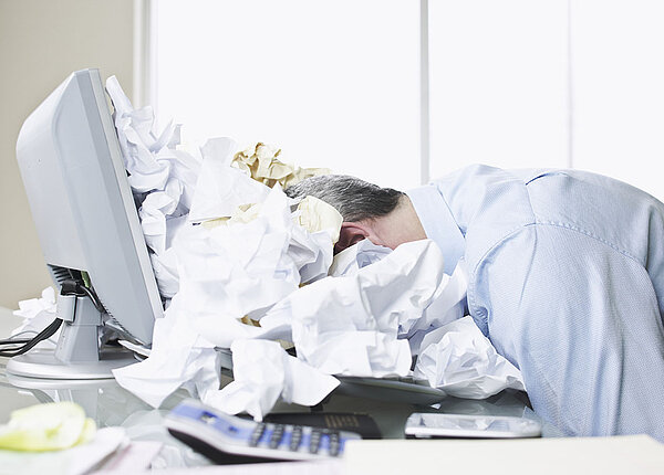 Tipps um Stress am Arbeitsplatz vorzubeugen Mann liegt mit seinem Kopf auf seinem Laptop in einem Meer aus zerknülltem Papier