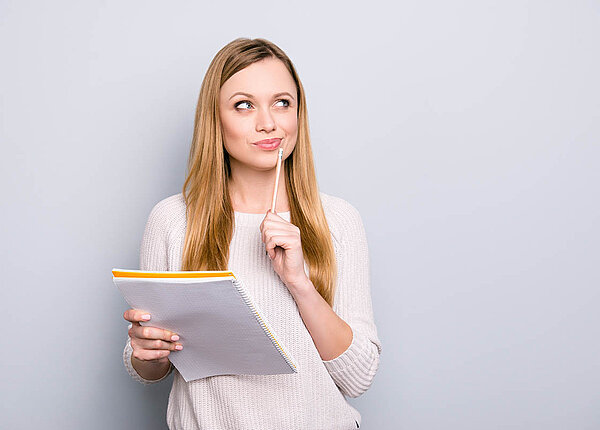 Vorgetäuschte Produktivität: Wie die Arbeitskultur dazu beiträgt und wie Authentizität bei der Arbeit wieder gelingen kann. Eine Frau hält in der einen Hand einen Block, mit der anderen hält sie sich einen Stift an den Mundwirkel, sie grübelt grinsend.