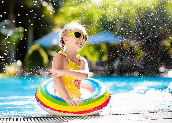 Infektionen und Folgen der Sonne beim Schwimmen in der Hitze plus Empfehlungen Mädchen trägt eine gelbe Herz-Sonnenbrille, einen gelben Badeanzug und einen Regenbogen-Schwimmreif. Sie steht am Schwimmbeckenrand und spritzt mit Wasser.