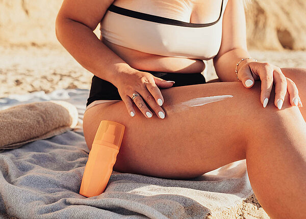 Sonnencreme und Umwelt: Infos zu mineralischen UV-Filtern, korallenfreundlicher Sonnencreme und Mikroplastik in den Meeren Eine Frau sitzt am Strand auf ihrem Handtuch. Sie cremt sich den Oberschenkel ein, an ihr Bein ist eine Flasche Sonnencreme gelehnt.