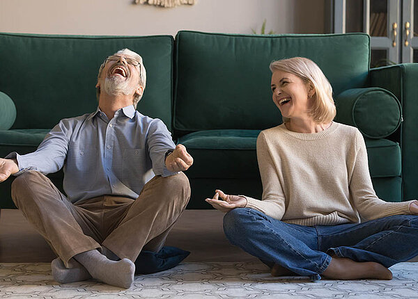 Lachen als Therapie: Atemübungen helfen bei COPD. Ein älteres Paar sitzt zum Meditieren auf dem Wohnzimmerboden und muss lachen.