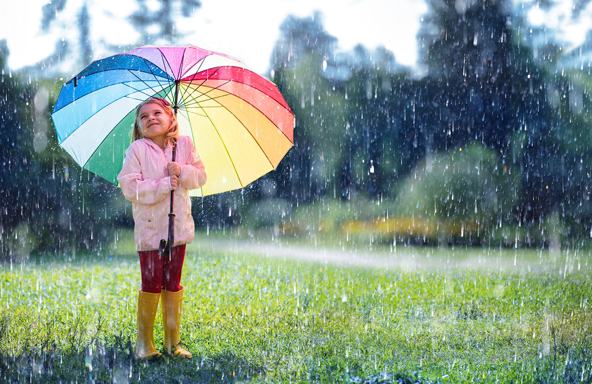 Ein Mädchen im Vorschulalter steht im Regen auf einer Wiese, schützt sich mit einem bunten Schirm vor den Tropfen und lächelt.