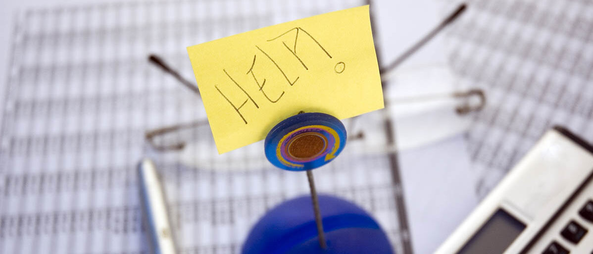 Auf mehreren ausgedruckten Tabellen liegen ein Taschenrechner, ein Stift, eine Brille sowie ein Schild mit der Aufschrift „HELP!“