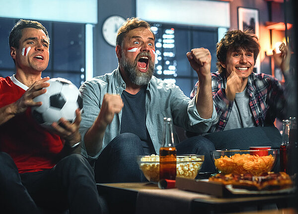 Drei Freunde sitzen auf einem Sofa und schauen ein Fußballspiel im TV. Sie jubeln.