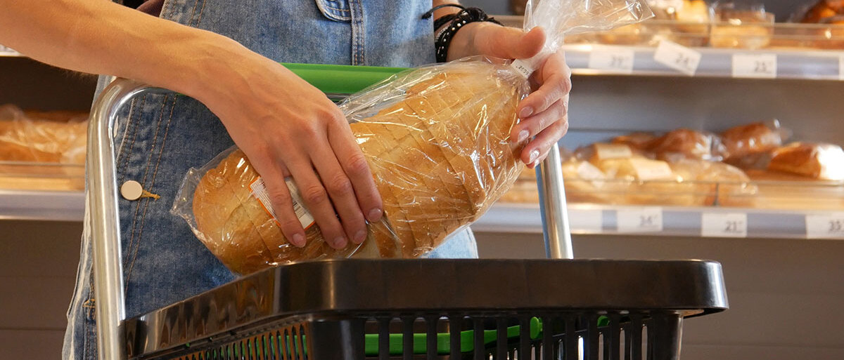 Eine Frau in einer Latzhose legt ein geschnittenes, abgepacktes Weißbrot in ihren Einkaufskorb.