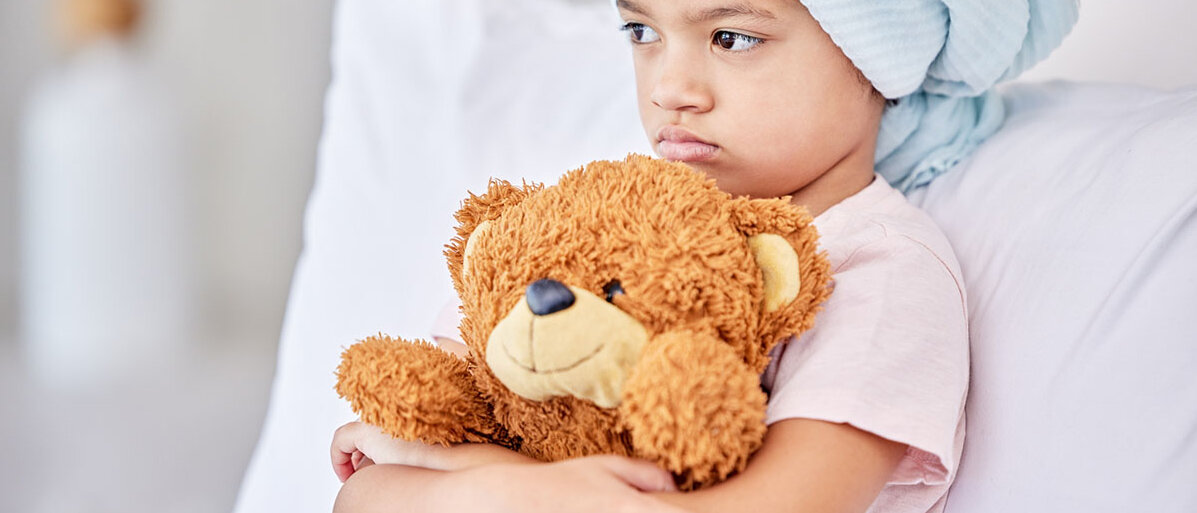 Ein Mädchen im Grundschulalter sitzt angelehnt in einem Krankenhausbett. Sie trägt ein Tuch auf dem Kopf, kuschelt ihren Teddy und schaut mit nach unten gezogenen Mundwinkeln ins Leere.