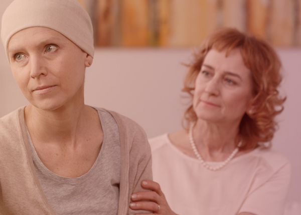 Krebsbehandlung und Nebenwirkungen von Krebsmedikamenten: Allgemeine Beratungstipps rund um die Therapie Eine Krebspatientin mit Mütze sitzt auf dem Sofa und schaut in die Ferne. Eine weitere Frau sitzt hinter ihr, schaut sie an und hält ihren Arm.