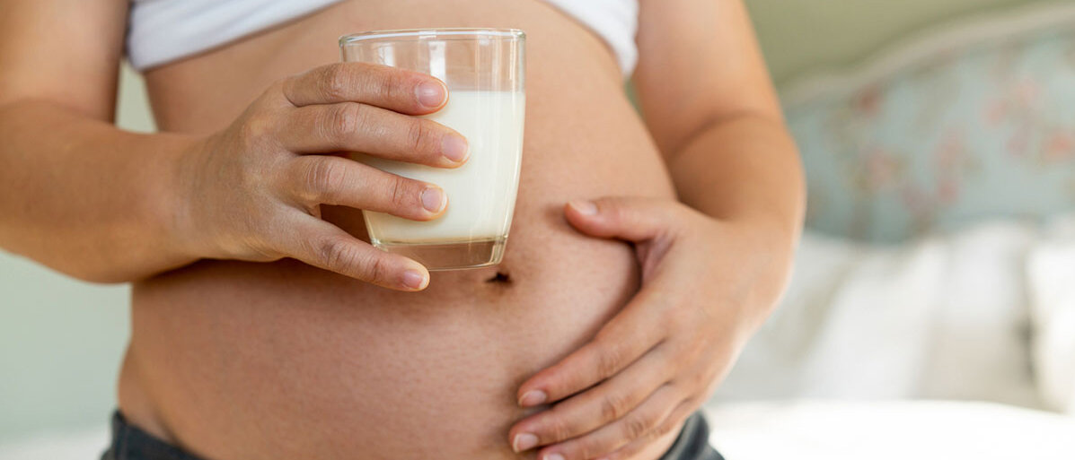 Schwangeren Bauch. Frau haelt in einer Hand ein Glas Milch und die andere Hand liegt auf dem Bauch.