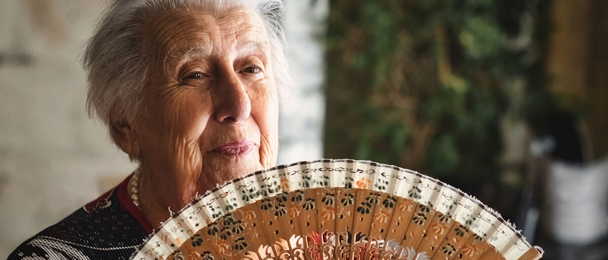 Eine alte Frau sitzt im Schatten und fächelt sich mit einem Fächer Luft zu.