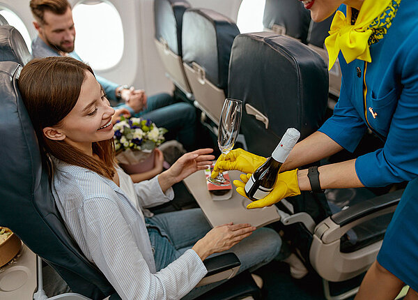 Eine junge Frau sitzt auf dem Gangplatz eines Flugzeugs. Die Stewardess reicht ihr ein Sektglas und einen Piccolo.