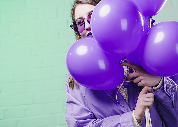 Junge Frau mit einer lila Sonnenbrille, lila Übergangsjacke und lila Lippenstift, die hinter einem Bündel lila Luftballons hervorblickt. 