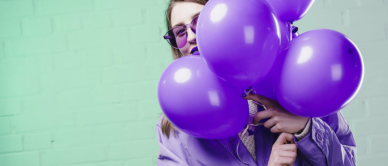 Junge Frau mit einer lila Sonnenbrille, lila Übergangsjacke und lila Lippenstift, die hinter einem Bündel lila Luftballons hervorblickt. 