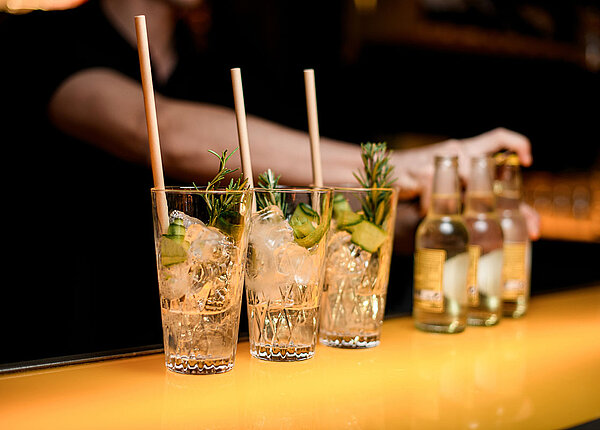 Auf einer Bar stehen drei Longdrink-Gläser mit Gin, Eis, Gurke und Rosmarin. Der Barkeeper öffnet gerade drei Fläschchen Tonic Water, die daneben stehen.