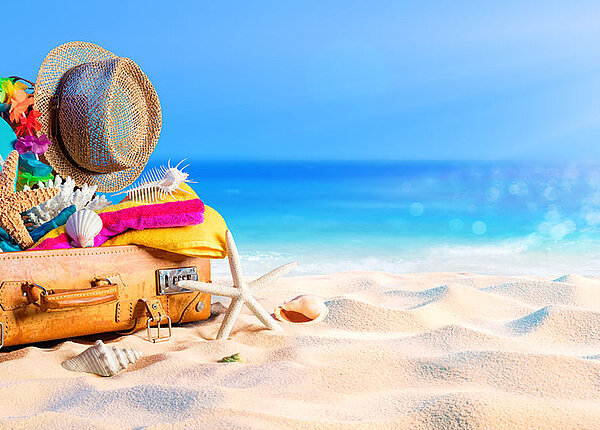 aufgeklappter altmodischer Koffer, indem zahlreiche Urlaubsutensilien stecken, darunter Flip-Flops, ein Handtuch, eine Sonnenbrille und ein Sonnenspray. Außerdem im und um den Koffer befinden sich Seesterne und Muscheln. Er liegt auf Sand, im Hintergrund das Meer.
