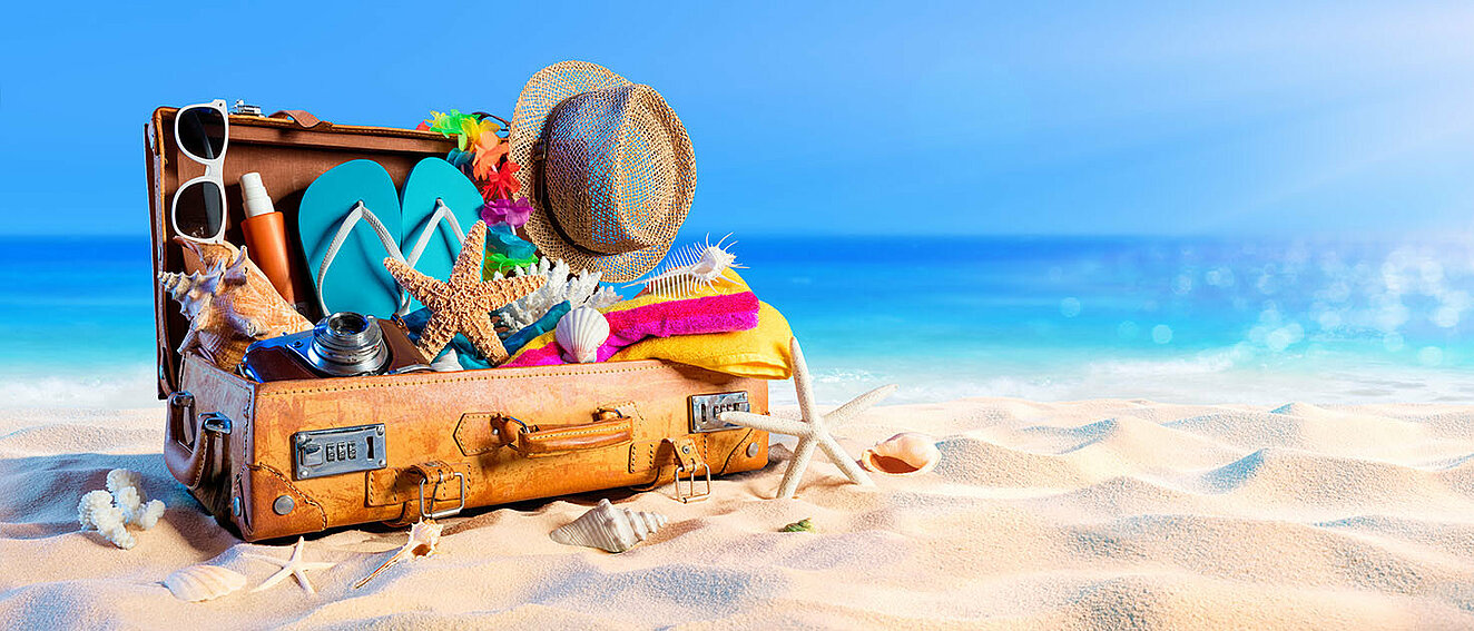 aufgeklappter altmodischer Koffer, indem zahlreiche Urlaubsutensilien stecken, darunter Flip-Flops, ein Handtuch, eine Sonnenbrille und ein Sonnenspray. Außerdem im und um den Koffer befinden sich Seesterne und Muscheln. Er liegt auf Sand, im Hintergrund das Meer.