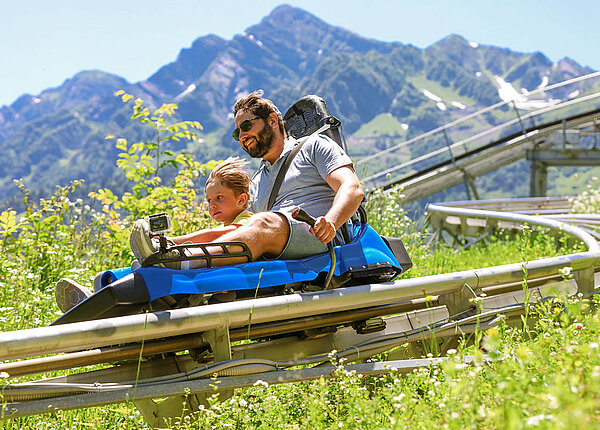 Mama und kleiner Junge sitzen in einem blauen Rodelschlitten und fahren eine Rodelbahn herunter. Im Hintergrund sind die Berge zu sehen.