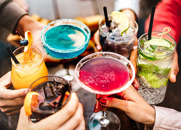 Sechs Menschen prosten sich mit verschiedenen bunten Cocktails in unterschiedlichen Gläsern zu.