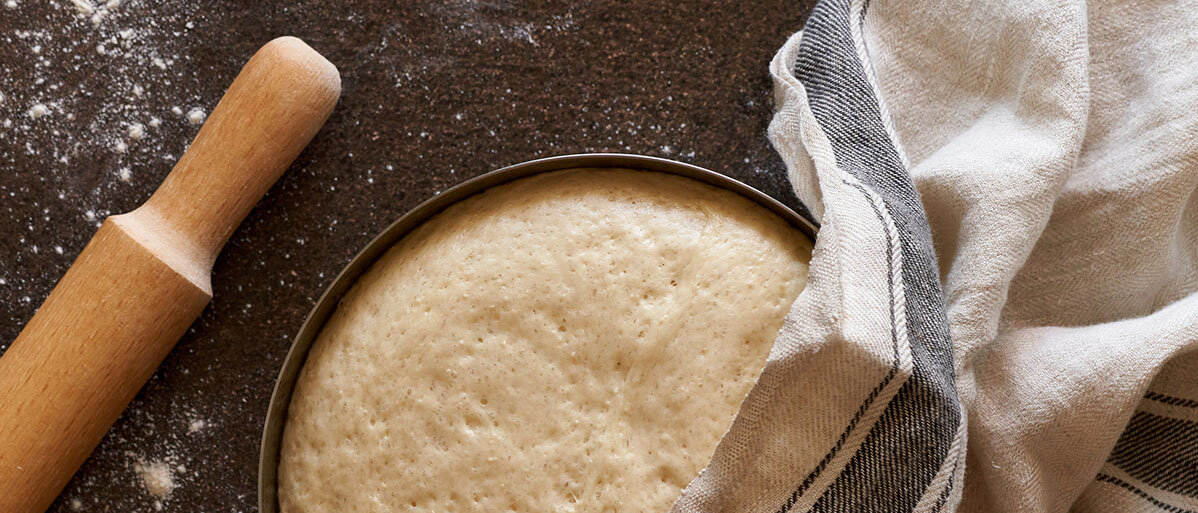 Hefeteig richtig einfrieren und auftauen Roher Hefeteig in einer Schüssel leicht bedeckt von einem Küchenhandtuch. Nebendran liegt ein Nudelholz.