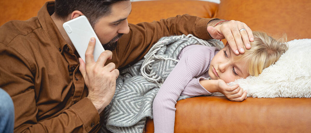 Ein Mädchen im Grundschulalter liegt erschöpft auf dem Sofa. Der Vater befühlt die Stirn, mit der anderen Hand telefoniert er.