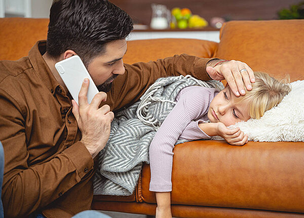 Ein Mädchen im Grundschulalter liegt erschöpft auf dem Sofa. Der Vater befühlt die Stirn, mit der anderen Hand telefoniert er.