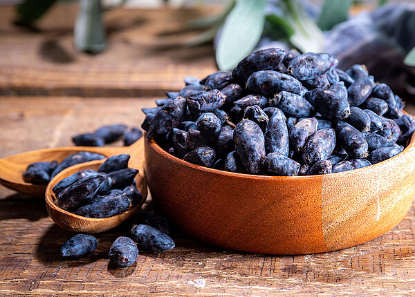 blaue Beeren in einer brauner Schuessel. Links daneben liegen zwei Holzlöffel und auf der vorderen liegen ebenfalls blaue Beeren