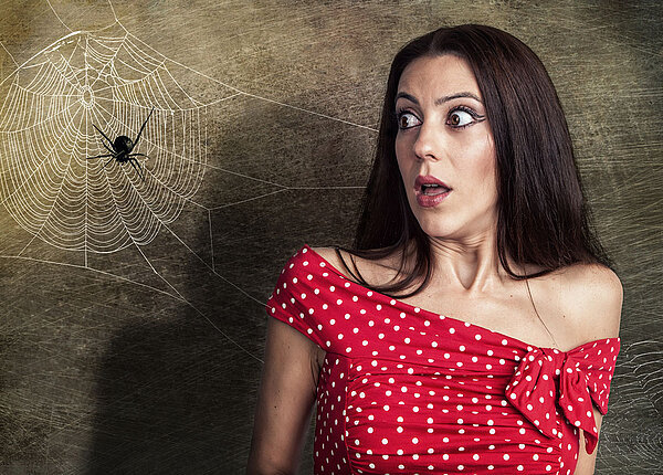 Frau mit einem roten Kleid mit weißen Punkten steht neben einem Spinnennetz, auf der eine Spinne zu sehen ist und sie hat Angst