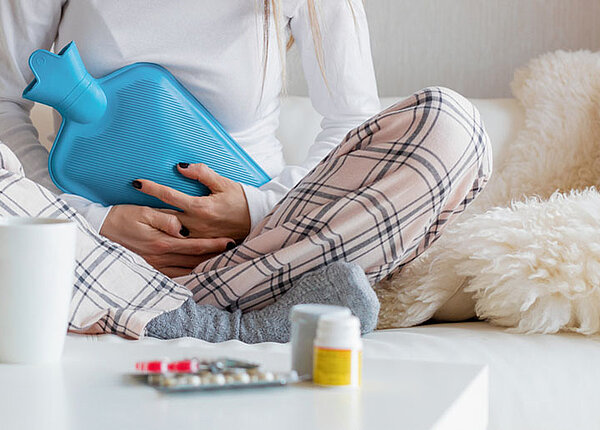 Frau sitzt im Pyjama und mit einer Wärmflasche auf dem Bauch auf der Couch. Vor ihr Medikamente gegen Regelschmerzen.