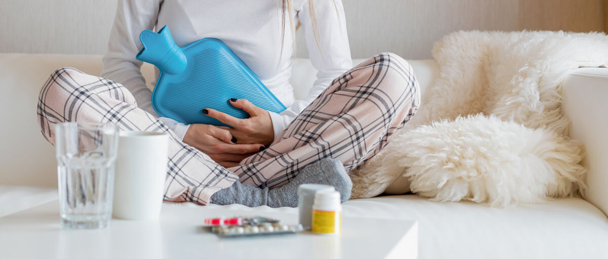 Frau sitzt im Pyjama und mit einer Wärmflasche auf dem Bauch auf der Couch. Vor ihr Medikamente gegen Regelschmerzen.