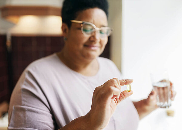 Eine Frau hält in der einen Hand ein Glas Wasser, in der anderen Hand hält sie eine oblonge, transparente Weichkapsel zwischen Daumen und Zeigefinger. Sie schaut auf die Kapsel und lächelt.