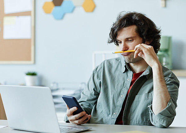 Junger Mann sitzt vor Laptop und hält einen Bleistift unter der Nase und schaut auf sein Handy.