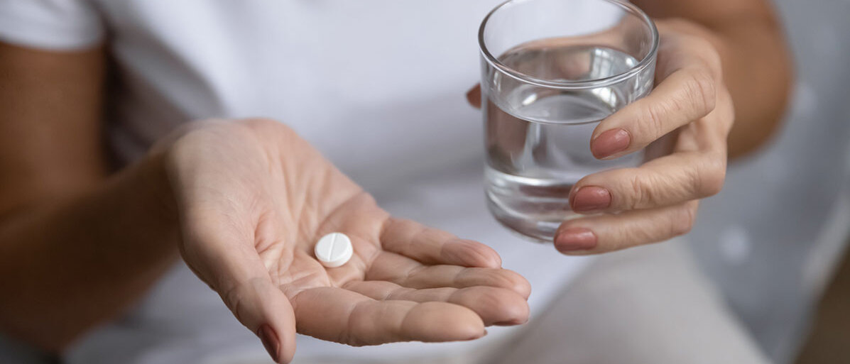 In einer Frauenhand liegt eine weiße Tablette und die andere Hand hält ein Wasserglas.