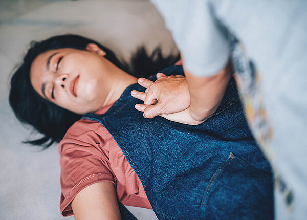 Erste Hilfe Maßnahmen: Notruf, Herzdruckmassage und wie man richtig hilft Eine Frau liegt regungslos und mit geschlossenen Augen auf dem Boden. Eine andere Frau gibt ihr mit verschränkten Händen eine Herzdruckmassage.