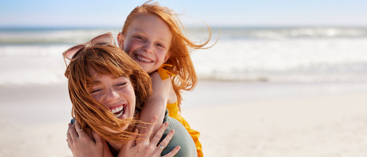 Mädchen umarmt ihre Mutter von hinten und beide lächeln. Beide sind am Strand