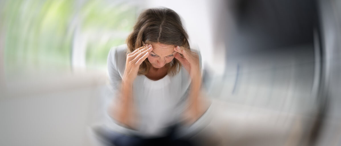 Sitzende Frau mit dunklen Haaren hat ihre Hände an den Schläfen und um se herum dreht sich alles.