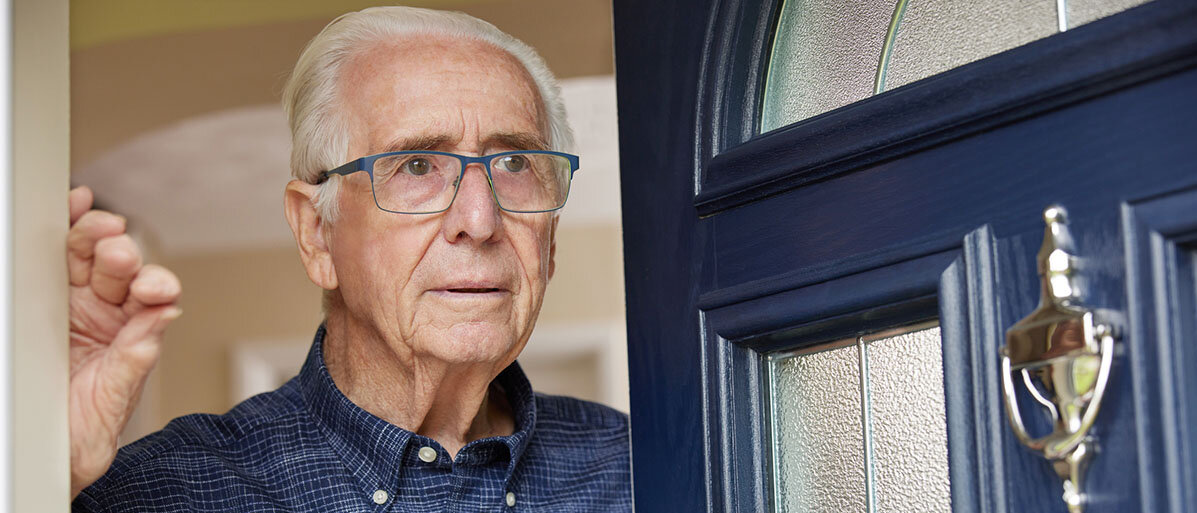 älterer Mann mit Brille steht an seiner blauen Wohnungstür.