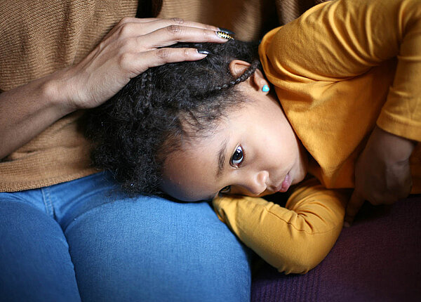 Antibiotika fürs Kind: Ist danach eine Darmsanierung mit Probiotika nötig, um das Darmmikrobiom zu stabilisieren? Ein kleines Mädchen liegt mit dem Kopf auf dem Schoß seiner Mutter, schaut erschöpft und hält eine Hand an den Bauch.