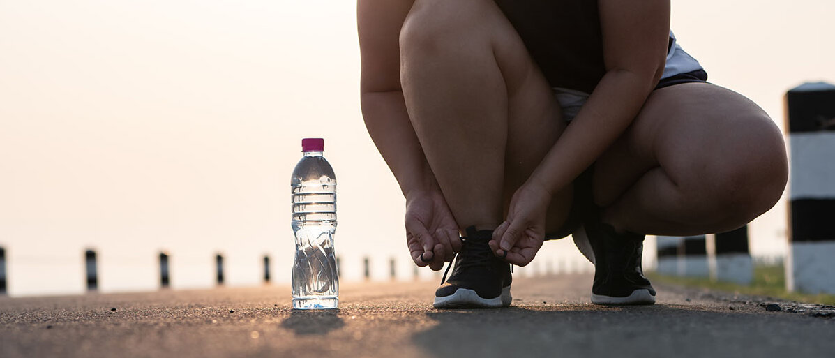 Eine Joggerin kniet sich in der Dämmerung hin, um ihre Laufschuhe zu binden. Die tiefstehende Sonne scheint durch ihre Wasserflasche.