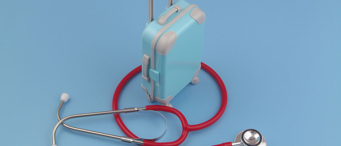Reisekoffer, drum herum liegt ein rotes Stethoskop auf blauem Hintergrund