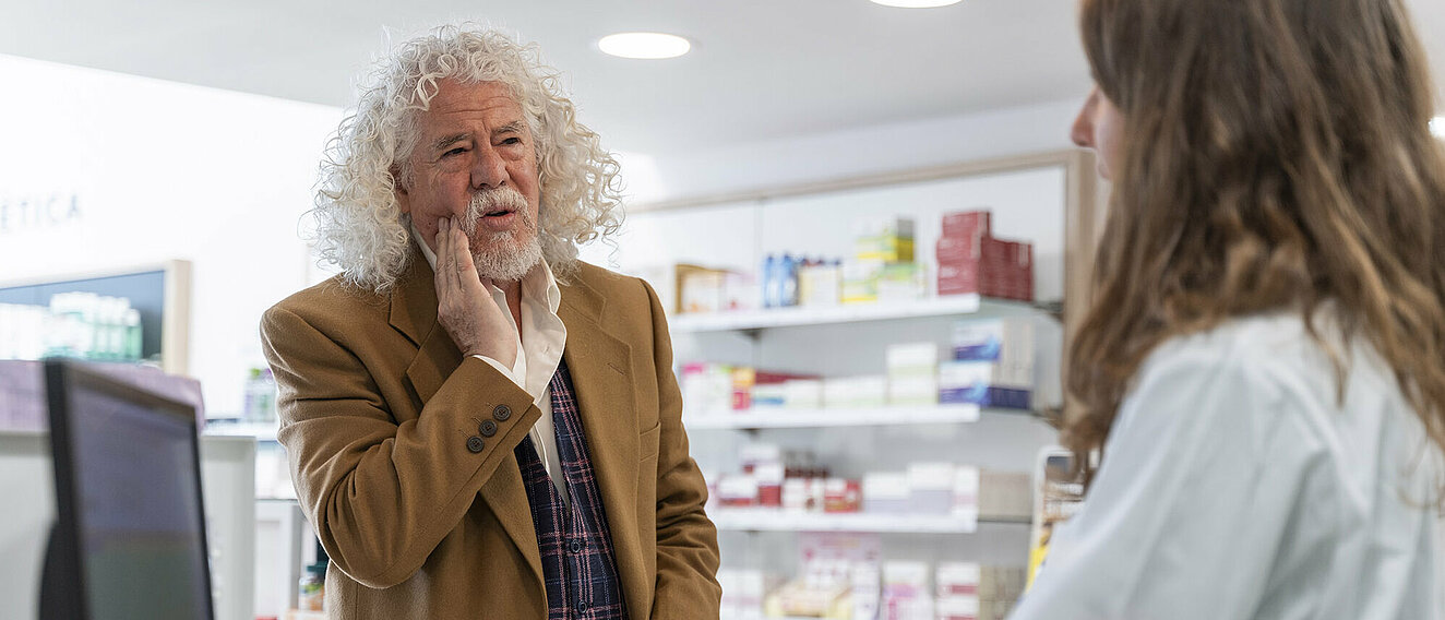 Ein älterer Herr in der Apotheke im Beratungsgespräch mit einer jungen PTA oder Apothekerin. Er hält sich die Wange.