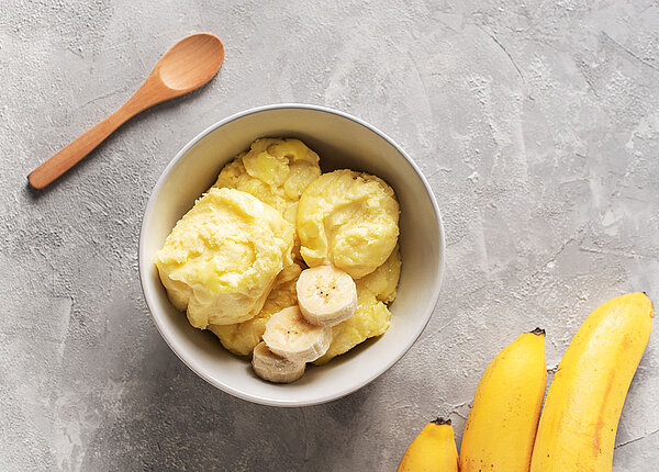 Ein Becher mit Bananeneis, daneben ein Löffel aus Holz und drei Bananen