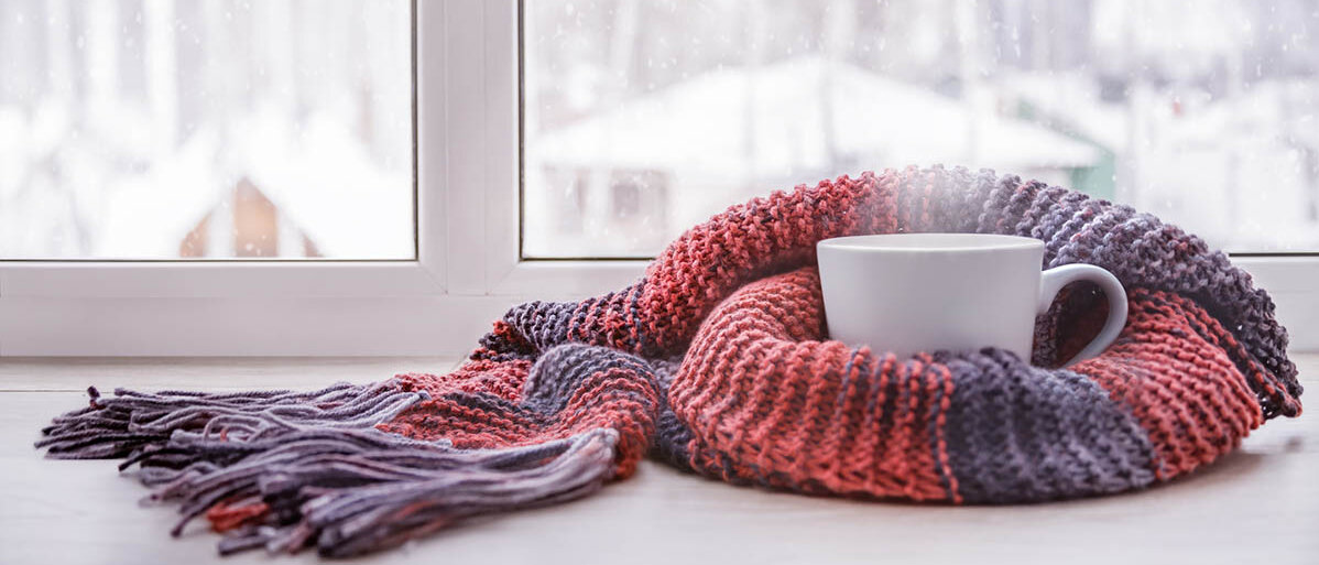 Erkältung: Husten und Halsschmerz Auf einer Fensterbank steht eine dampfende Tasse, um die ein Strickschal geschlungen ist. Draußen liegt Schnee.