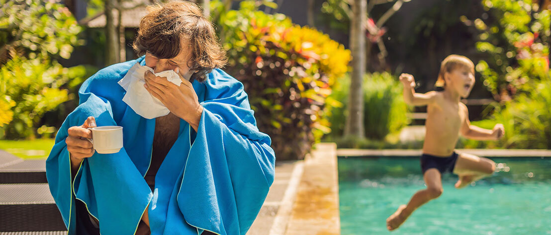 Vater uns Sohn am Pool. Der Sohn springt ins Wasser, während sich der Vater - sichtlich krank - die Nase putzt